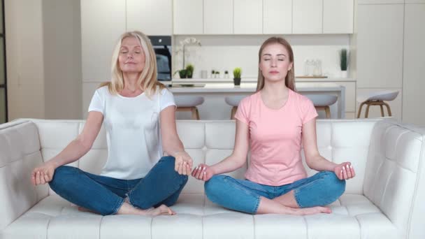 Feliz familia madre mayor e hija adulta en el sofá practicando meditación de yoga en casa. Mujer adulta y su madre mayor haciendo yoga felizmente juntas para meditar. — Vídeo de stock
