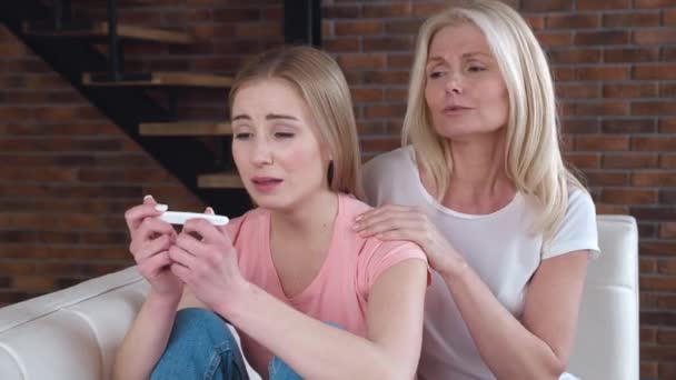 Young adult woman is suffering and crying from an unwanted pregnancy, holding a pregnancy test in her arms, sitting on the couch at home. Senior Mom Calms Her Adult Daughter With Parental Support — Stock Video