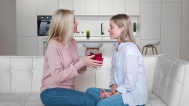 Kıdemli anne, mutlu yetişkin kızına doğum günü hediyesi kutusunu sunuyor gülümseyen, kucaklayan, olgun bir kadın evdeki kanepede yetişkin kızını kucaklıyor.