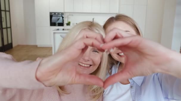 Gelukkig glimlachende oudere moeder en haar volwassen dochter maken hartvorm handgebaar, zittend op de bank thuis. Familie liefde concept — Stockvideo