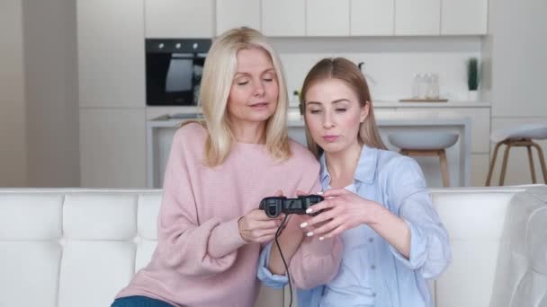 Счастливая взрослая дочь учит свою старшую мать играть в видеоигры на консоли, сидя на диване перед телевизором. Два поколения женщин семьи проводят весело время вместе дома. — стоковое видео