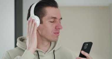 Genç bir adamın yakın plan portresi. Beyaz kulaklıklarla müzik dinliyor, akıllı bir telefonla kafasını evin içinde ritimde sallıyor.
