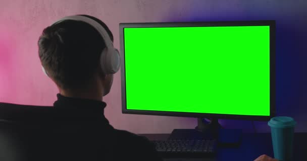 Achteraanzicht van een jongeman met koptelefoon kijkend op monitor met groene chroma toets, binnen. Achteraanzicht van mannelijke student kijkt video, op een computer monitor in de avond thuis. — Stockvideo