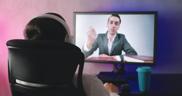 वीडियो ब्लॉग, इनडोर देखने वाले हेडफ़ोन वाले एक युवा के पीछे का दृश्य। पुरुष छात्र का बैक व्यू एक प्रशिक्षण वीडियो, घर पर शाम को कंप्यूटर मॉनिटर पर एक ऑनलाइन वेबिनार देखता है . — स्टॉक वीडियो