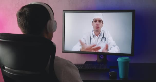 Vista trasera de un hombre joven viendo video conferencia en línea con el médico. Solo paciente hombre está consultando con el médico en el chat en línea en la noche. — Vídeos de Stock