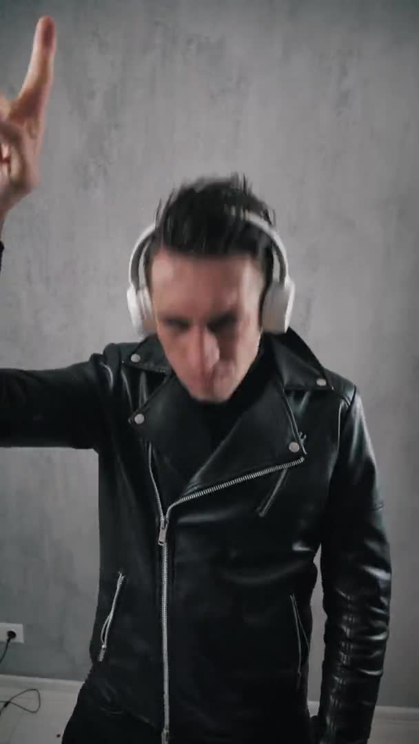 Vertical video of man rocker in leather jacket enjoying heavy rock and roll music with headphones and swinging to the rhythm showing goat gesture — Stock Video