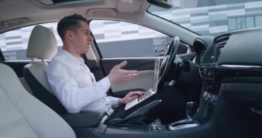 Arabada otururken bilgisayarla konuşan genç iş adamının portresi. Erkek girişimci görüntülü sohbet yoluyla selamlıyor, uzaktan çalışma konsepti.