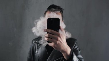 Deri ceketli sigara içen adam yüzünü akıllı telefonla gizler ve koyu gri arka planda duman üfler.