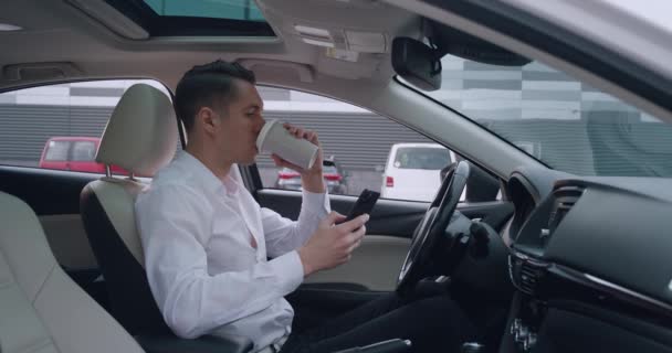 Retrato de jovem empresário senta-se no banco dos motoristas em um carro e usa smartphone para navegar nas redes sociais e beber café. — Vídeo de Stock