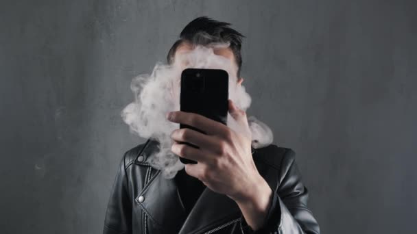 Smoker man in leather jacket hide his face with smartphone and blowing smoke over dark grey background — Stock Video