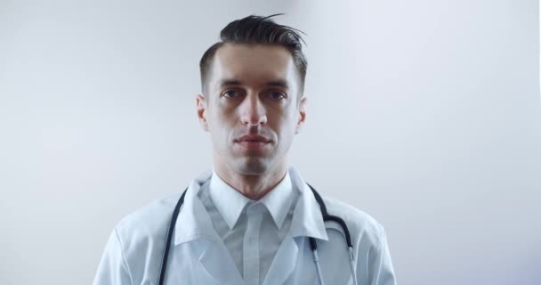 Porträt eines männlichen Arztes, der eine medizinische Maske aufsetzt und vor weißem Hintergrund in die Kamera blickt. Vorsichtsmaßnahmen gegen Grippe und Coronavirus — Stockvideo
