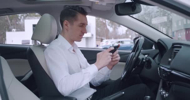 Homem de negócios de camisa branca excitadamente joga um jogo em seu smartphone enquanto sentado no banco dos motoristas no carro. — Vídeo de Stock