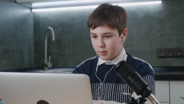 Bonito menino saudação e bate-papo on-line por videochamada com computador portátil e microfone profissional em casa, enquanto sentado à mesa. Comunicação à distância — Vídeo de Stock