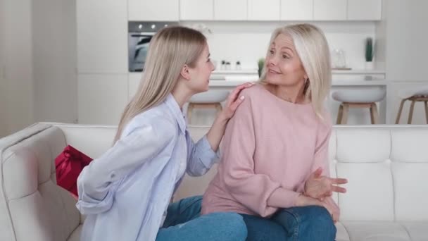 Adult daughter presenting birthday gift box to happy old mother smiling bonding embracing, young woman congratulating hugging senior mom on mothers day on sofa at home — Stock Video
