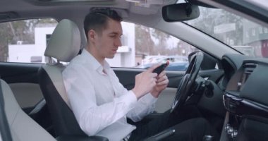 Arabada sürücü koltuğunda otururken beyaz gömlekli gülümseyen genç adam akıllı telefonuyla uygulama oyunu oynuyor..
