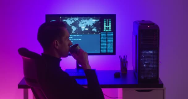 Mužský programátor pije kávu, když pracuje v noci u počítače v datovém centru. Nebezpečný hacker pracující v centru kybernetické bezpečnosti naplněné neonovým světlem. — Stock video