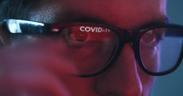 メガネに反映されたコンピュータで働いている男のクローズアップ、 COVID 19の警告。若い男性は夜にインターネット上でコロナウイルスのニュースを閲覧します. — ストック動画
