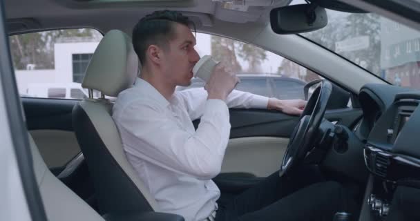Beyaz gömlekli genç işadamının portresi arabada oturup kahve içiyor. Yakışıklı erkek sürücü kameraya bakıyor. — Stok video