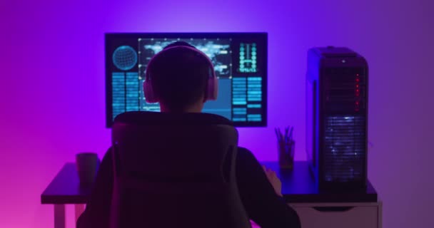 Programista w słuchawkach pracujący w nocy przy komputerze w centrum danych. Niebezpieczny haker pracujący w centrum cyberbezpieczeństwa wypełnionym światłem neonowym. — Wideo stockowe