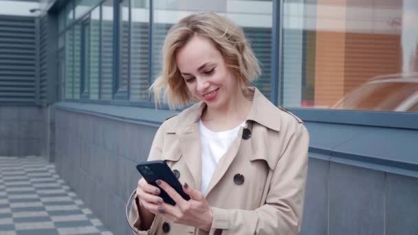 Portret van een glimlachende jonge vrouw in een regenjas met smartphone, genieten van surfen op sociale netwerken, buiten staan op een moderne kantoorachtergrond — Stockvideo