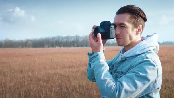 Mladý fotograf fotografující na profesionální fotokameře, stojící na červeném poli. Portrét muže používajícího digitální fotoaparát venku — Stock video