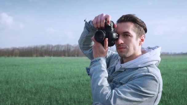 Jovem fotógrafo do sexo masculino tirando fotos em fotocâmera profissional, de pé no belo campo verde. Retrato de homem usando câmera digital ao ar livre — Vídeo de Stock