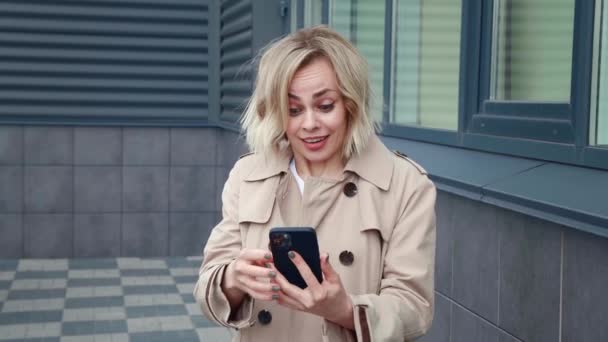 幸せなビジネス女性の肖像は、ダウンタウンの屋外に立っている間、スマートフォンでの成功をお楽しみください。若い女性は驚きましたすごいホールド使用して携帯電話,彼女の携帯電話で良いニュースを読む. — ストック動画