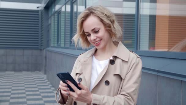Ritratto di giovane donna felice sorridente in impermeabile con smartphone, godendo di navigare sui social network, in piedi all'aperto su sfondo moderno edificio per uffici — Video Stock