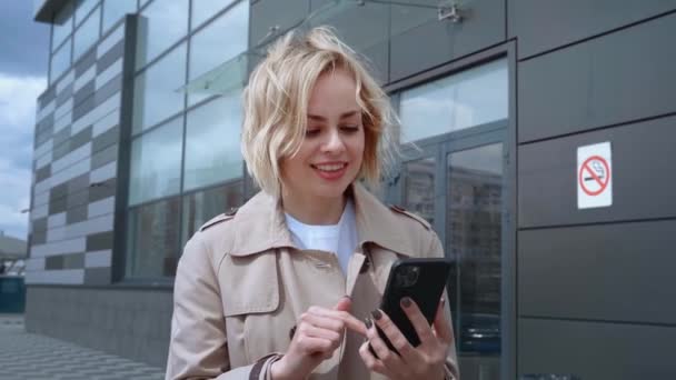 Retrato de una joven mujer de negocios en una chaqueta caminando por la ciudad y mensajes de texto en el teléfono inteligente. Mujer rubia bonita usando el teléfono móvil mientras camina al aire libre. — Vídeo de stock