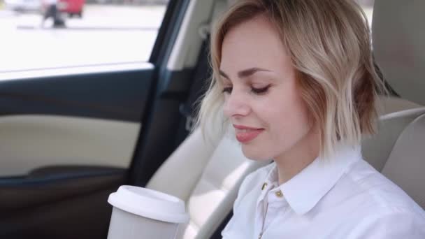 Retrato de una mujer de negocios sonriente con camisa blanca sentada en el coche y tomando café. Conductora bastante femenina mirando a la cámara — Vídeo de stock