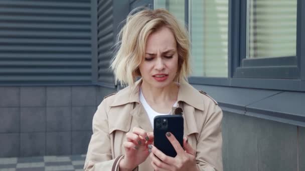 Upset business woman looking with horror at the smartphone screen and covers her mouth with her hand in fear. Surprised caucasian woman reading terrible news on mobile phone while stands outdoors — Stock Video