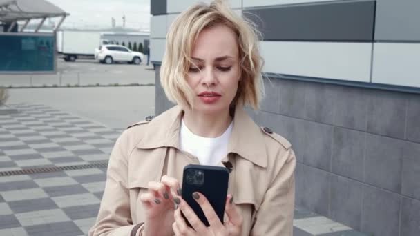 Портрет молодої бізнес-леді в куртці, що гуляє по місту і текстових повідомленнях на смартфоні. Красива блондинка-жінка використовує мобільний телефон під час прогулянки на відкритому повітрі . — стокове відео