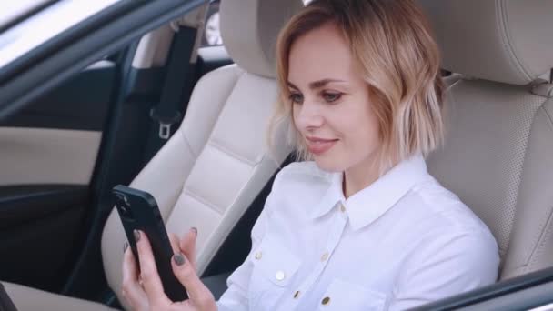 Hermosa mujer joven sentada en un coche en el asiento del conductor y el uso de teléfono inteligente. Mujer de negocios sonriente sosteniendo el teléfono en el coche. — Vídeos de Stock