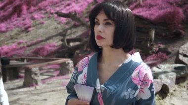 Japon geyşası gibi giyinmiş güzel beyaz bir kadının sakura ağaçlarının arkasında dikilirken çekilmiş bir fotoğrafı.