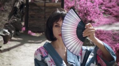 Geleneksel Japon kimonolu beyaz geyşa kadının yakın çekimi, yüzünü ahşap el vantilatörüyle kapatıyor ve açık havada kırmızı sakura ağaçlarının arkasında duruyor. Sabit çekim