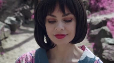 Japon geyşası gibi giyinmiş güzel beyaz bir kadının sakura ağaçlarının arkasında dikilirken çekilmiş bir fotoğrafı.