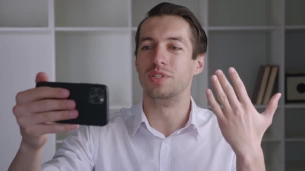 Retrato de hombre de negocios feliz haciendo videollamadas en tiempo real con teléfono inteligente en la oficina moderna, saludando a la gente en la pantalla del teléfono. Joven macho haciendo selfie chat de vídeo en línea en el teléfono móvil — Vídeo de stock