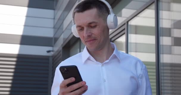 Retrato de hombre de 40 años de edad disfrutar de escuchar música por auriculares inalámbricos y teléfonos inteligentes, de pie al aire libre. Sonriendo macho maduro disfrutar de la música con auriculares y teléfono móvil, fuera cerca del edificio de oficinas. — Vídeos de Stock