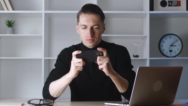 Fiatal férfi játszik az okostelefon izgalmas mobil alkalmazás megnyerte a versenyjáték szorító ököl örvendezik, miközben ül a munkahelyen az irodában. — Stock videók