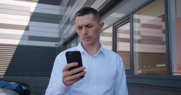 40 'lı yaşlarda beyaz tişörtlü bir erkek işadamı ofis binalarının yakınındaki cep telefonuna mesaj yolluyor. İş ve başarı kavramı — Stok video