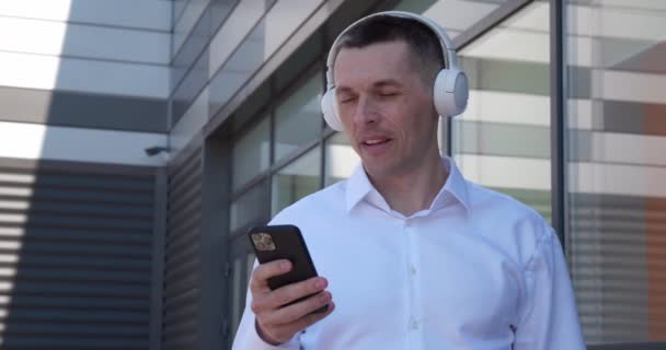 40 yaşındaki bir adamın portresi kablosuz kulaklıklar ve akıllı telefonlardan açık havada müzik dinlemekten hoşlanır. Gülümseyen olgun erkek, kulaklık ve cep telefonuyla ofis binasının önünde müzik dinlemekten hoşlanır.. — Stok video