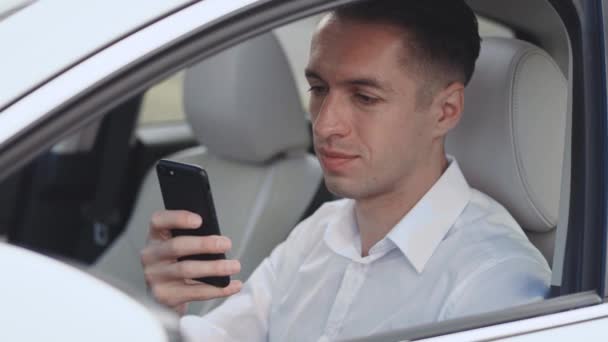 Крупним планом портрет усміхненого чоловічого бізнесмена, що сидить за кермом автомобіля і використовує смартфон . — стокове відео
