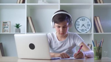 Kulaklıklı, kulaklıklı, bilgisayarındaki okul derslerine katılan, çocukluk çağındaki sevimli bir çocuk. Çünkü covid-19 ve not defterine notlar yazıyor. Uzaklık eğitimi çevrimiçi öğrenme dersi kavramı