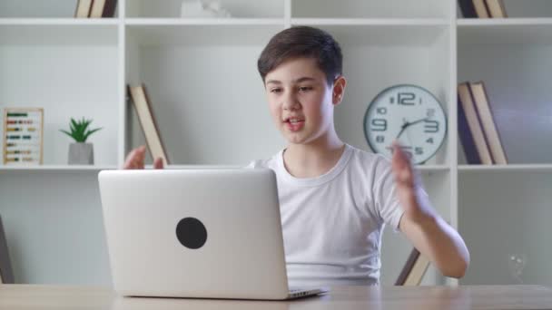 テーブルに座っている間、自宅でノートパソコンとのビデオ通話でオンラインチャットで笑顔の少年。家庭からのオンラインレッスンを見て学校の少年。距離学習 — ストック動画