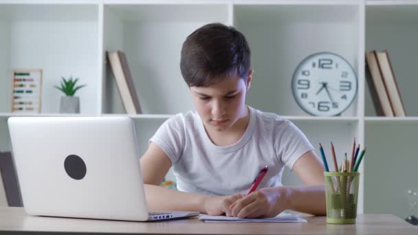 Молодой привлекательный мальчик-ученик посещает домашнюю онлайн-школу на своем ноутбуке из-за ковида-19 и пишет заметки на блокноте. Дистанционное обучение онлайн — стоковое видео