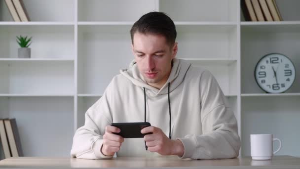 Νεαρός άνδρας παίζει στο smartphone του συναρπαστική εφαρμογή για κινητά κερδίζοντας ένα παιχνίδι αγώνων σφίγγοντας γροθιά χαρά, ενώ κάθεται στο χώρο εργασίας στο γραφείο. — Αρχείο Βίντεο