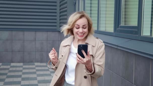 Portret van een gelukkige zakenvrouw vieren succes te winnen op de smartphone, buiten staan in het centrum. Jonge vrouw in regenjas verrast wow hold met behulp van mobiele telefoon, het lezen van goed nieuws op haar mobiele telefoon. — Stockvideo