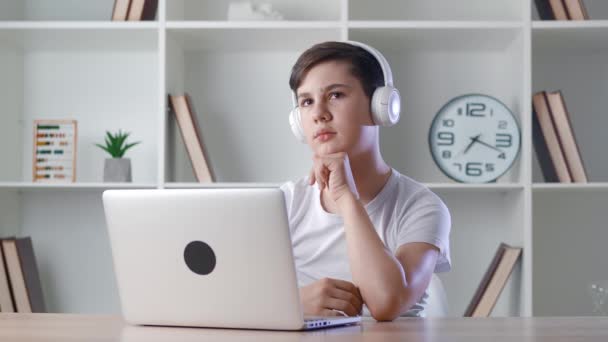 Pohledný zamyšlený školák 12-13 let ve sluchátkách, sedí u stolu a doma používá notebook. Mladý chlapec pracuje na notebooku na pozadí domácí kanceláře — Stock video