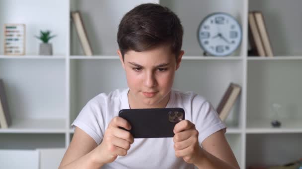 Όμορφο αγόρι 13 ετών που παίζει κινητό παιχνίδι στο smartphone στο σπίτι. Έφηβος παίζει κινητό τηλέφωνο. Το παιδί χρησιμοποιεί το τηλέφωνο για να παίξει. Παιδί που παίζει βιντεοπαιχνίδι σε εσωτερικούς χώρους — Αρχείο Βίντεο