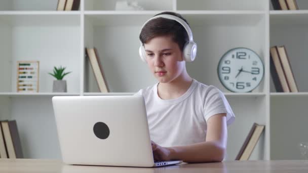 Hezký školák teenager 12-13 let ve sluchátkách, sedí u stolu a doma používá notebook. Mladý chlapec pracuje na notebooku na pozadí domácí kanceláře — Stock video